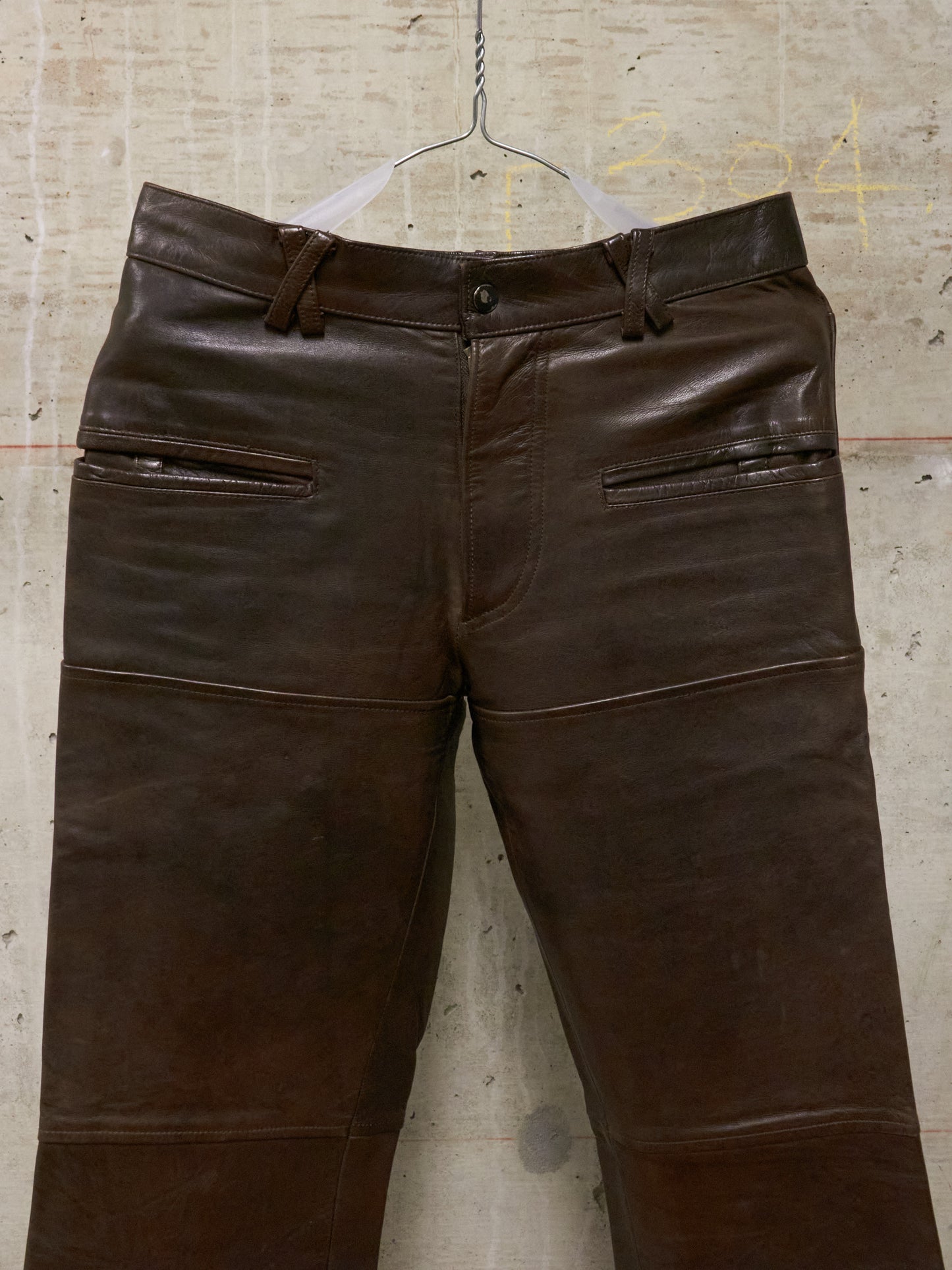 Brown leather pants on a hanger against a textured wall.