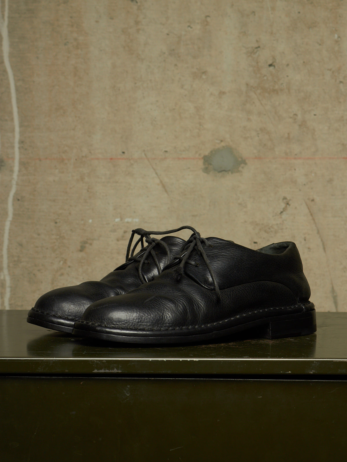 A pair of black leather lace-up shoes with a low block heel, displayed on a dark green surface against a textured beige concrete wall.