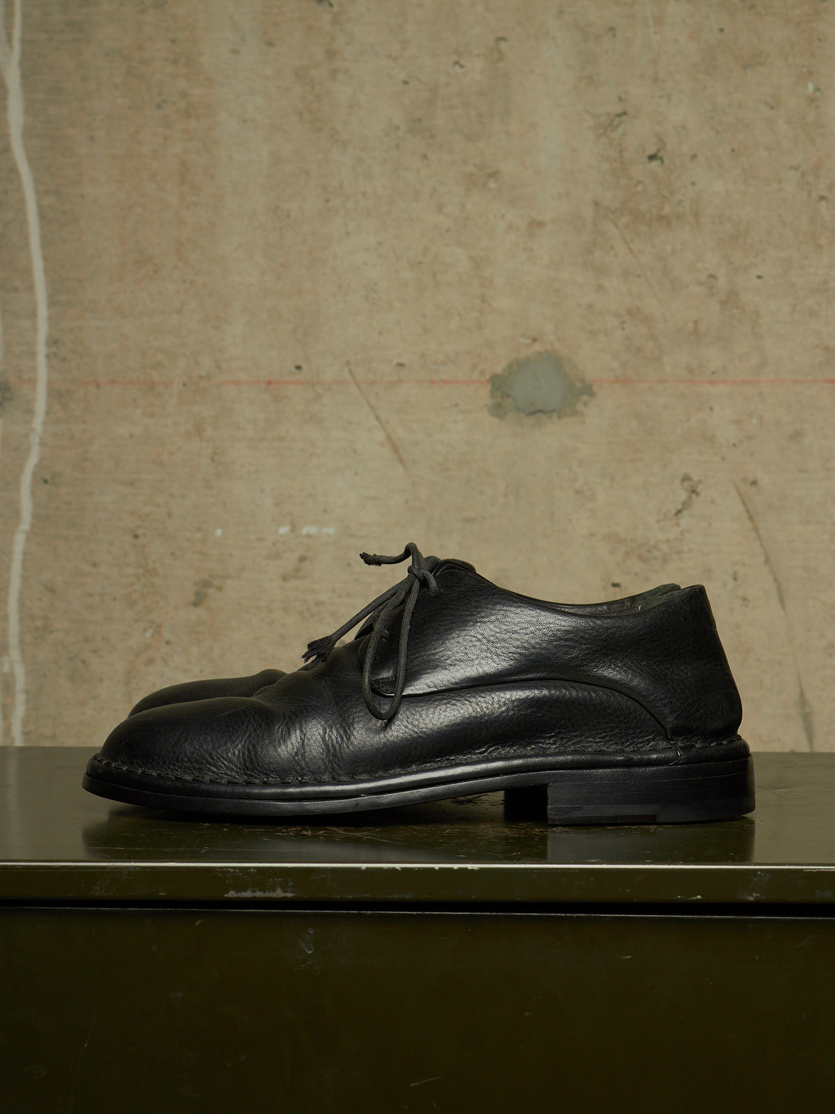 A pair of black leather lace-up shoes with a low block heel, displayed on a dark green surface against a textured concrete wall.