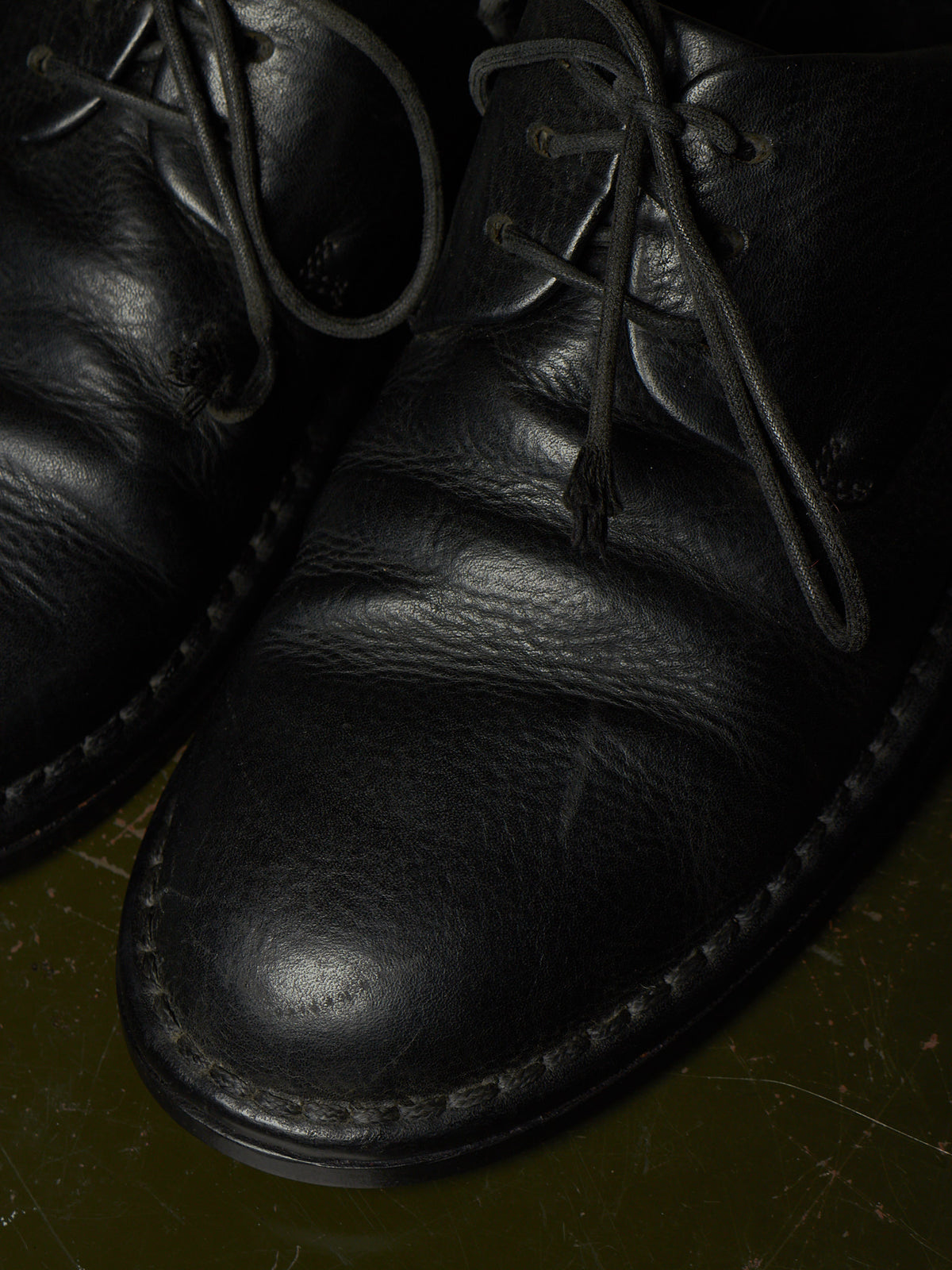 Close-up of a black leather lace-up shoe with visible grain texture and dark laces, photographed against a dark olive-toned background.