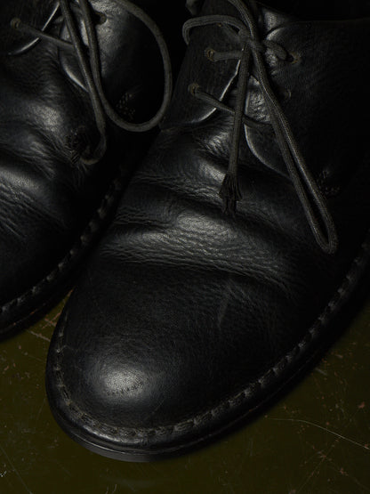 Close-up of a black leather lace-up shoe with visible grain texture and dark laces, photographed against a dark olive-toned background.