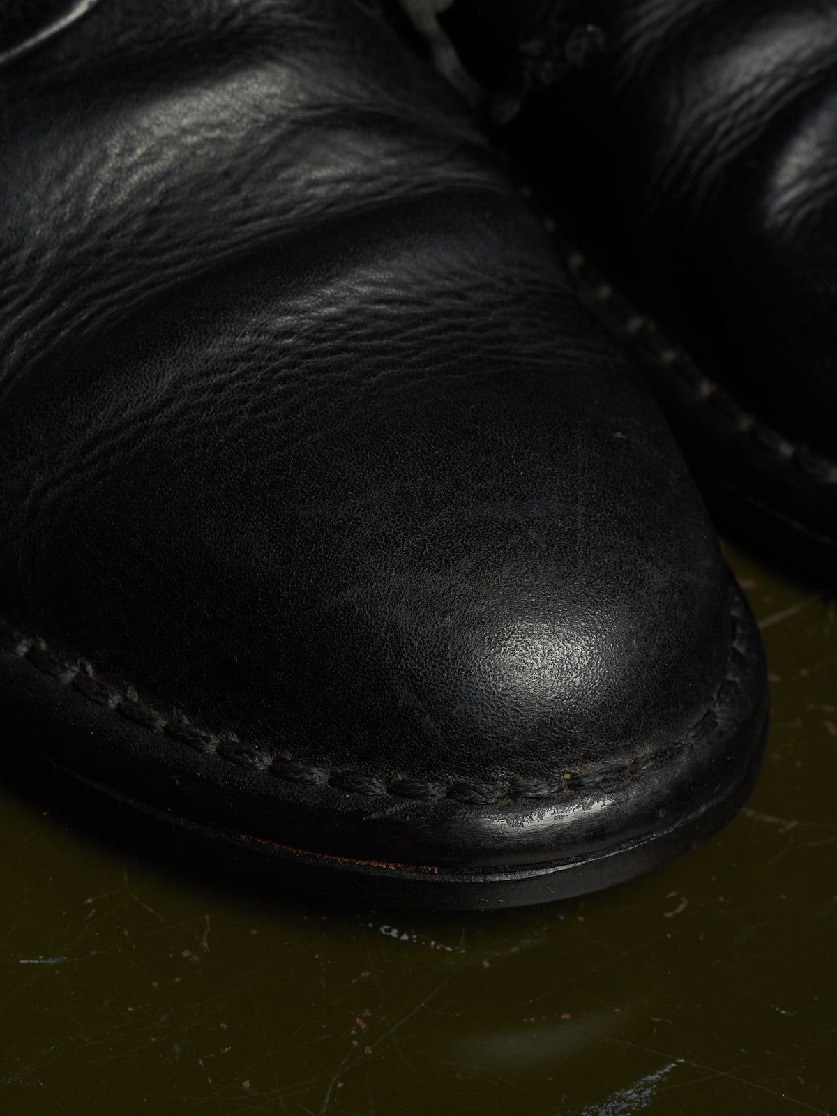 Close-up of a black leather boot toe with visible grain texture and contrast stitching, resting on a dark olive surface.