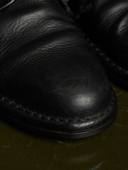Close-up of a black leather boot toe with visible grain texture and contrast stitching, resting on a dark olive surface.