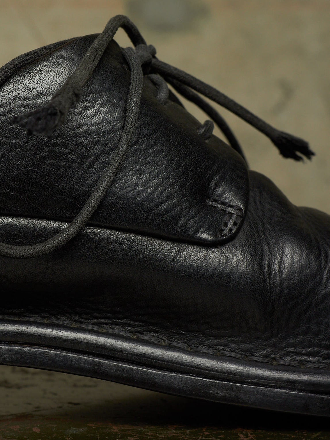 A close-up shot of a black leather lace-up shoe, showing the textured grain leather, stitching, and dark laces against a blurred neutral background.