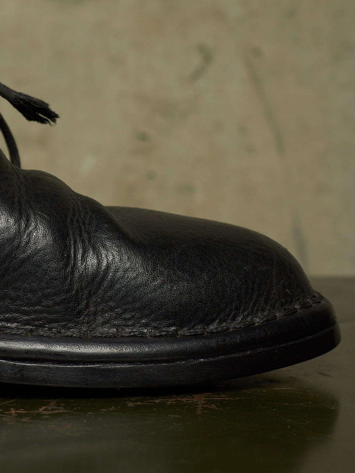 Close-up of a black leather boot toe with visible grain texture and black laces, resting on a dark surface against a warm beige background.