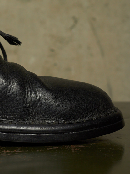 Close-up of a black leather boot toe with visible grain texture and black laces, resting on a dark surface against a warm beige background.
