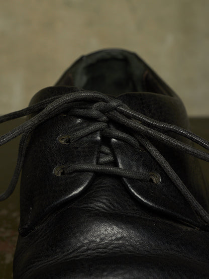A close-up shot of a black leather lace-up shoe, focusing on the textured leather and the tied laces against a neutral, out-of-focus background.