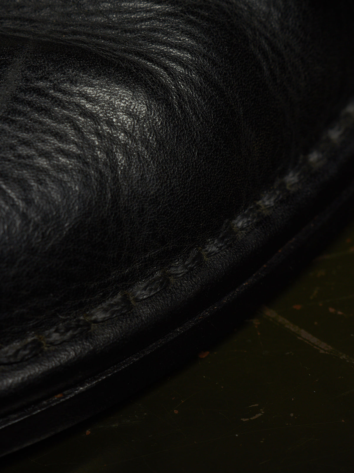 Extreme close-up of a black leather shoe or boot showing the grain texture and a stitched edge against a dark background.