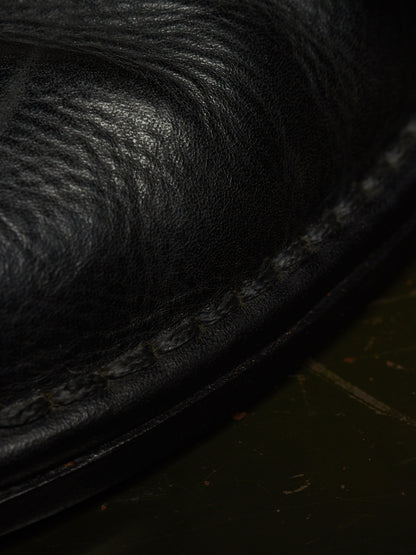 Extreme close-up of a black leather shoe or boot showing the grain texture and a stitched edge against a dark background.