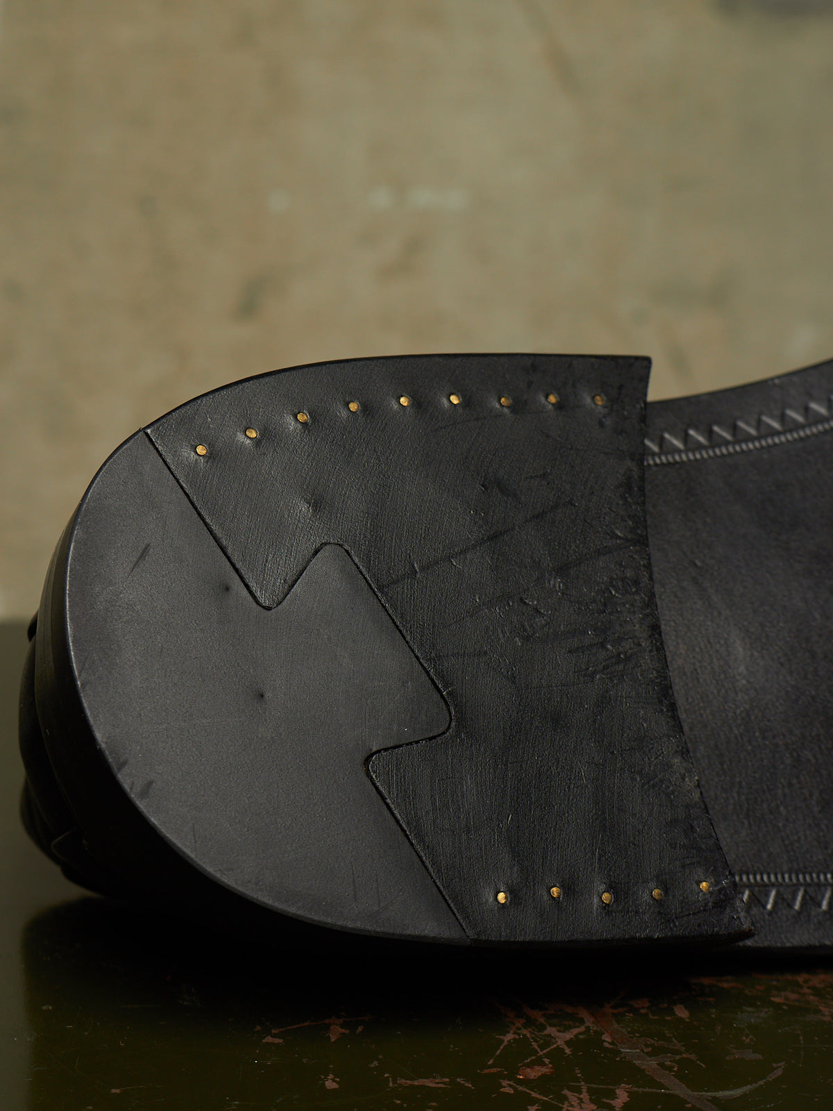 Close-up of the sole of a black leather shoe, showing stitched paneling and small brass-colored nailhead details against a textured neutral background.