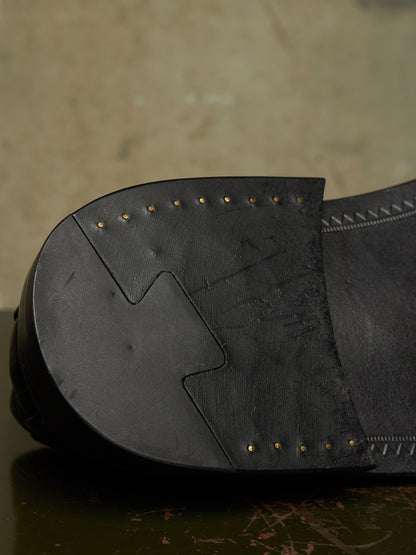 Close-up of the sole of a black leather shoe, showing stitched paneling and small brass-colored nailhead details against a textured neutral background.