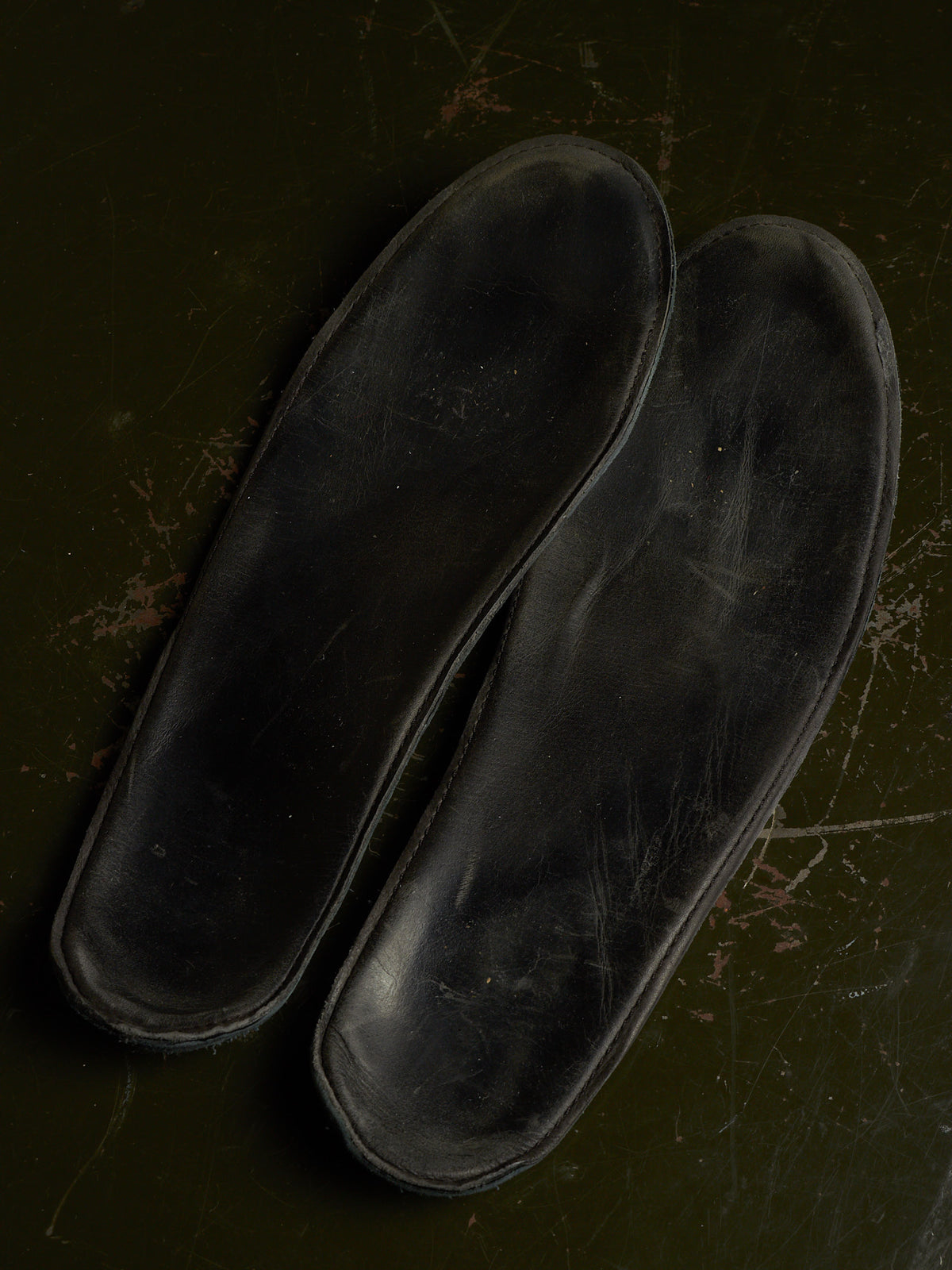 A pair of black shoe insoles photographed from above on a dark olive-green textured surface.