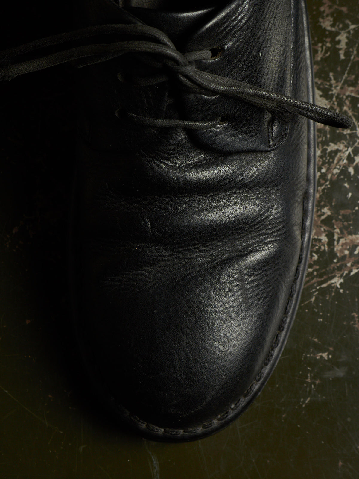 A close-up of a black leather lace-up shoe, showing the textured leather and laces, resting on a dark, worn surface.