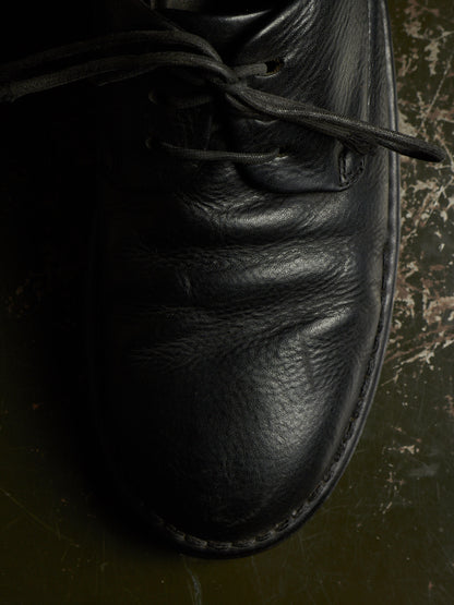 A close-up of a black leather lace-up shoe, showing the textured leather and laces, resting on a dark, worn surface.