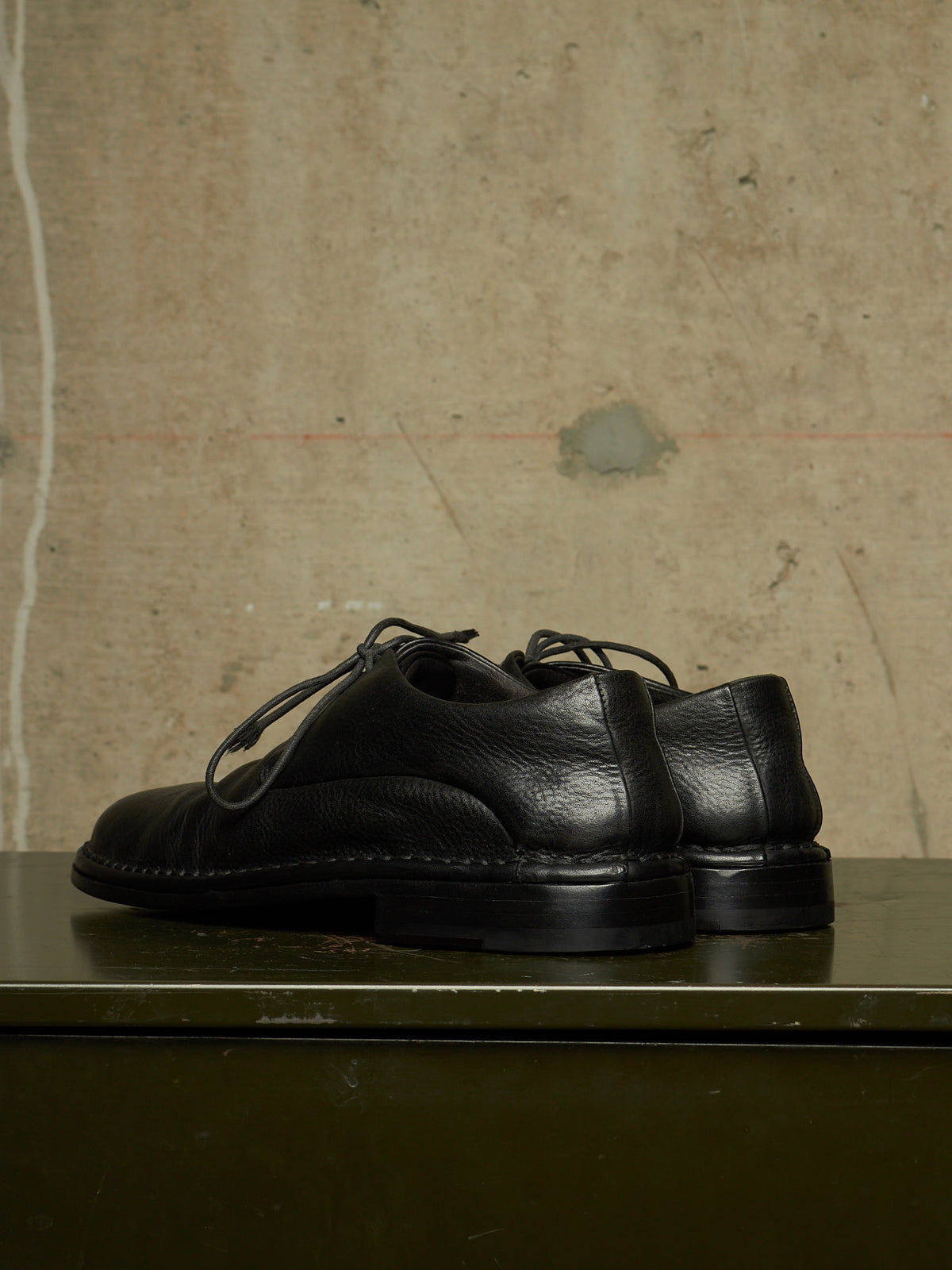 A pair of black leather lace-up shoes with a chunky sole, displayed on a dark green surface against a textured concrete wall.