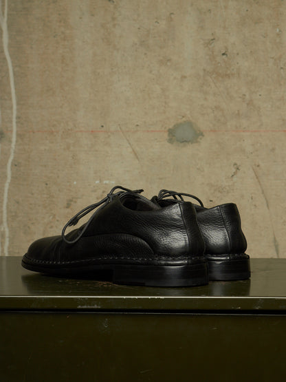 A pair of black leather lace-up shoes with a chunky sole, displayed on a dark green surface against a textured concrete wall.