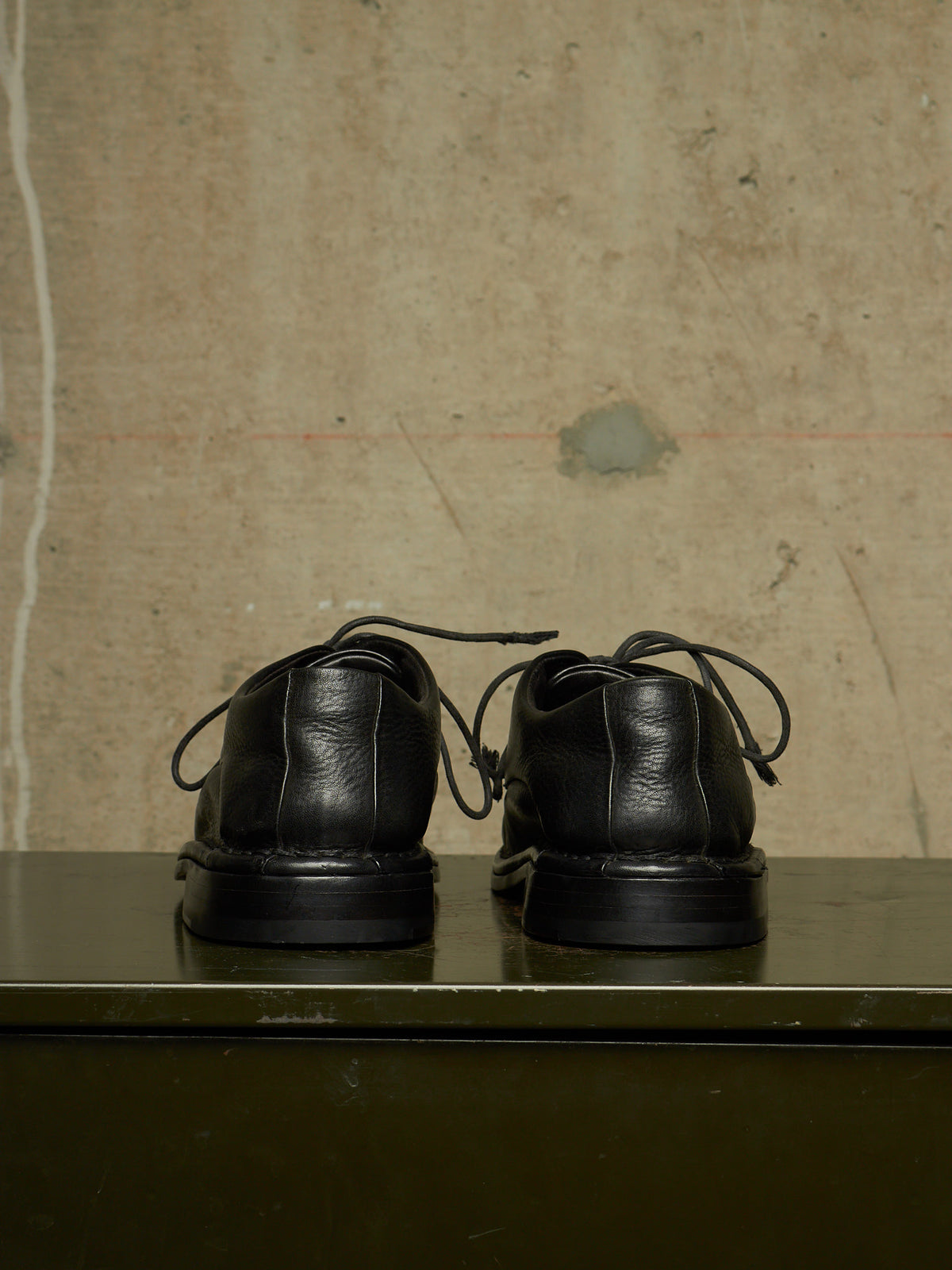 A pair of black leather lace-up shoes shown from the back, resting on a dark green surface against a textured beige concrete wall.