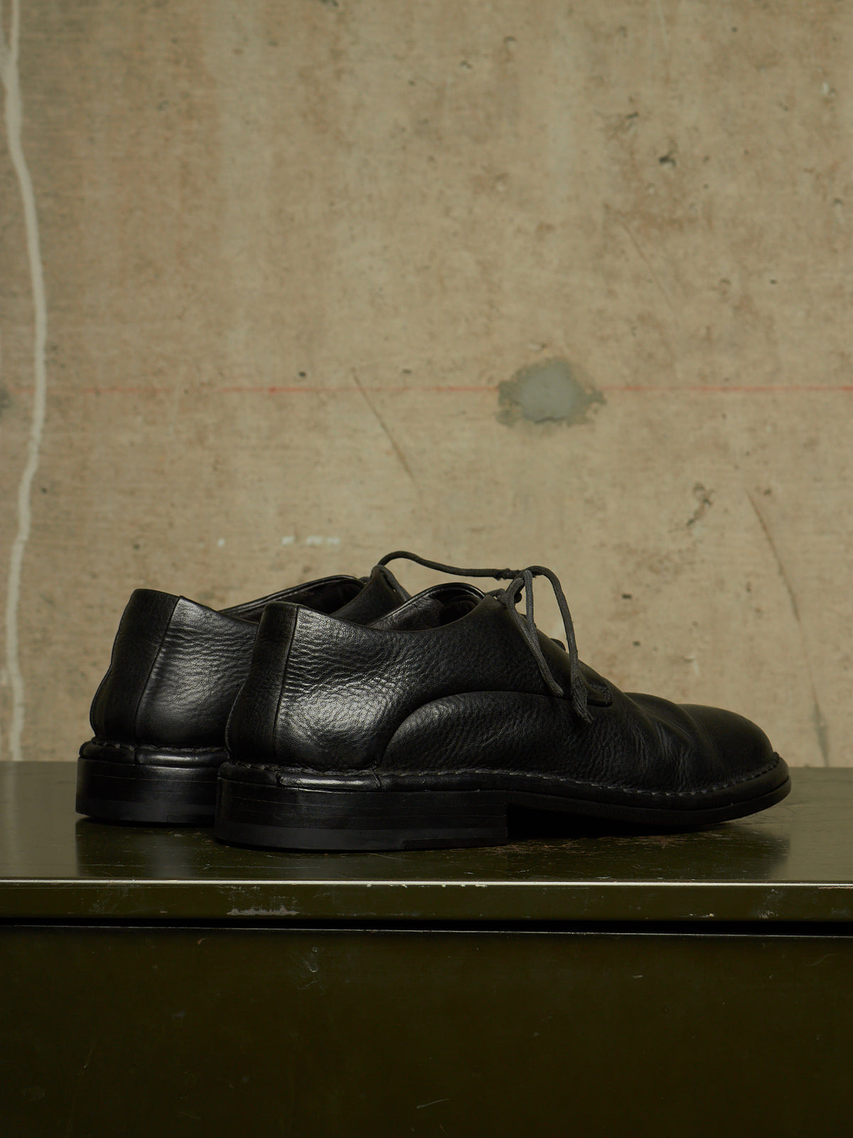 A pair of black leather lace-up dress shoes with a pebbled texture, resting on a dark green surface against a textured concrete wall background.