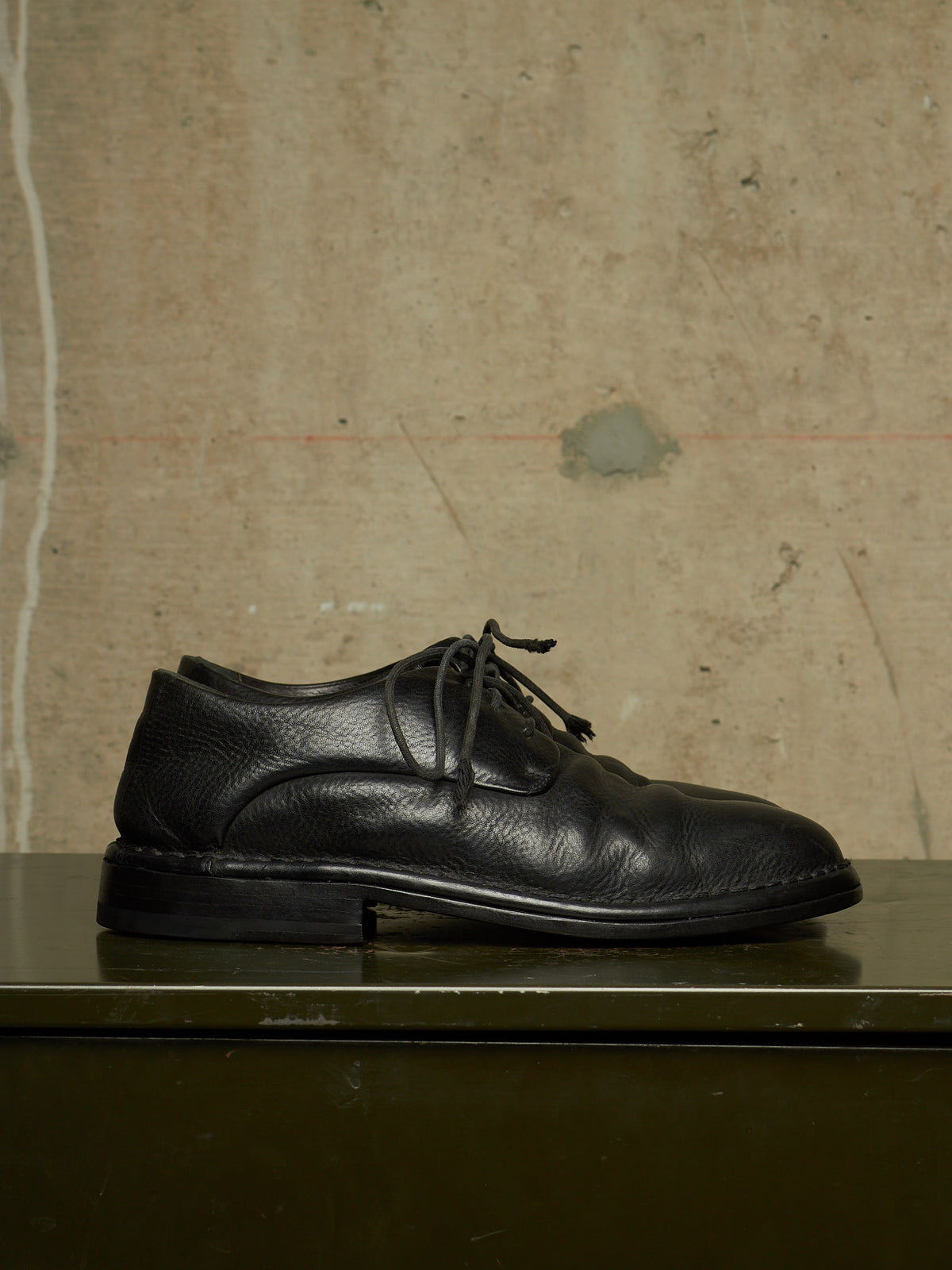 A single black leather lace-up dress shoe with a pebbled texture, resting on a dark green surface against a textured beige concrete wall.