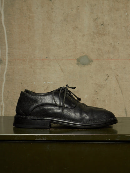 A single black leather lace-up dress shoe with a pebbled texture, resting on a dark green surface against a textured beige concrete wall.