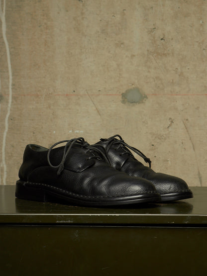 A pair of black leather lace-up dress shoes resting on a dark green surface against a textured, warm beige concrete wall.