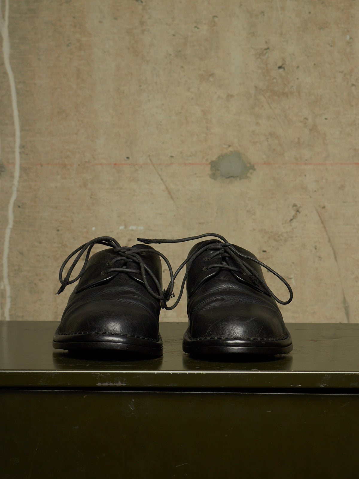 A pair of black leather lace-up shoes resting on a dark green surface against a textured beige concrete wall.