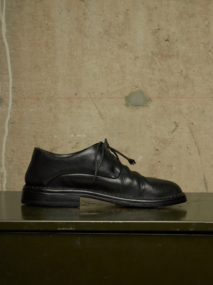 A single black leather lace-up dress shoe with a low block heel, resting on a dark green metal surface against a textured concrete wall background.