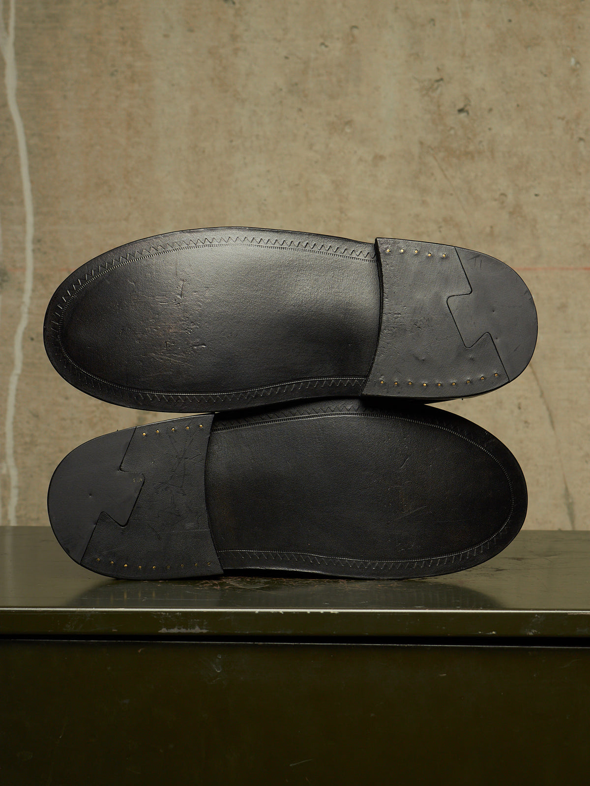 The soles of a pair of black leather loafers are shown from above, resting on a dark surface against a textured beige wall.