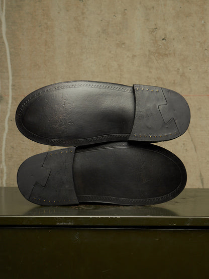 The soles of a pair of black leather loafers are shown from above, resting on a dark surface against a textured beige wall.