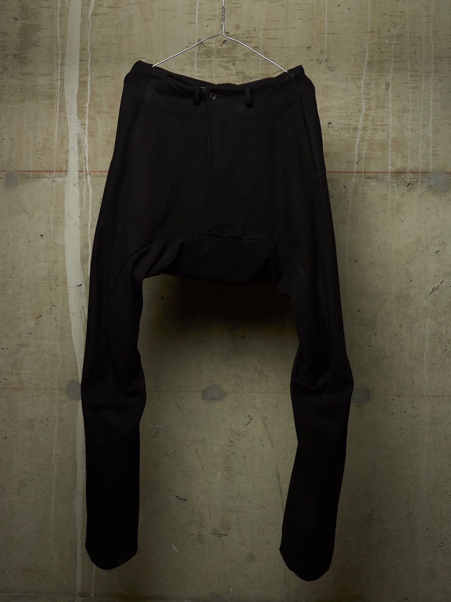 Black high-waisted, wide-leg trousers hanging from a metal wire hanger against a textured concrete wall.