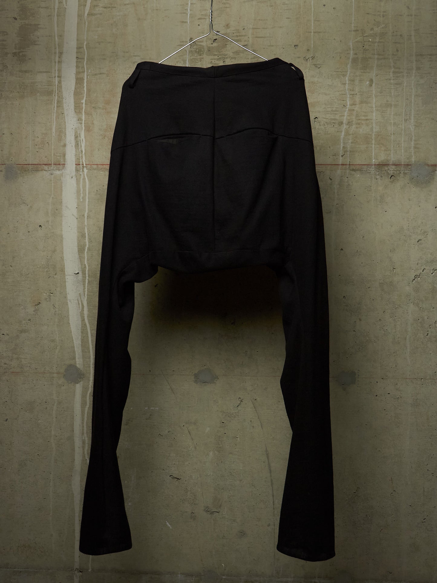 Black high-waisted cropped pants with a dramatic split leg design, hanging on a wire hanger against a textured concrete wall.