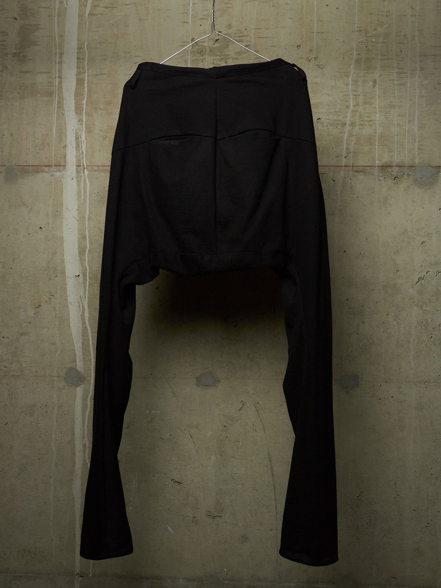 Black high-waisted cropped pants with a dramatic split leg design, hanging on a wire hanger against a textured concrete wall.