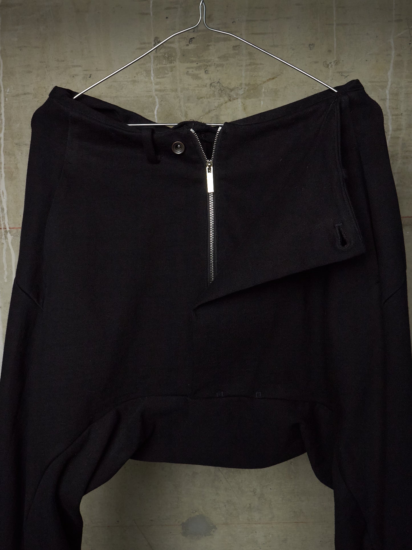Black asymmetrical draped pants with a front zipper and button closure, hanging on a metal hanger against a textured concrete wall.