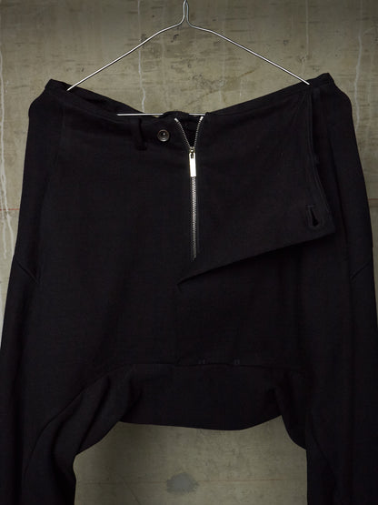Black asymmetrical draped pants with a front zipper and button closure, hanging on a metal hanger against a textured concrete wall.