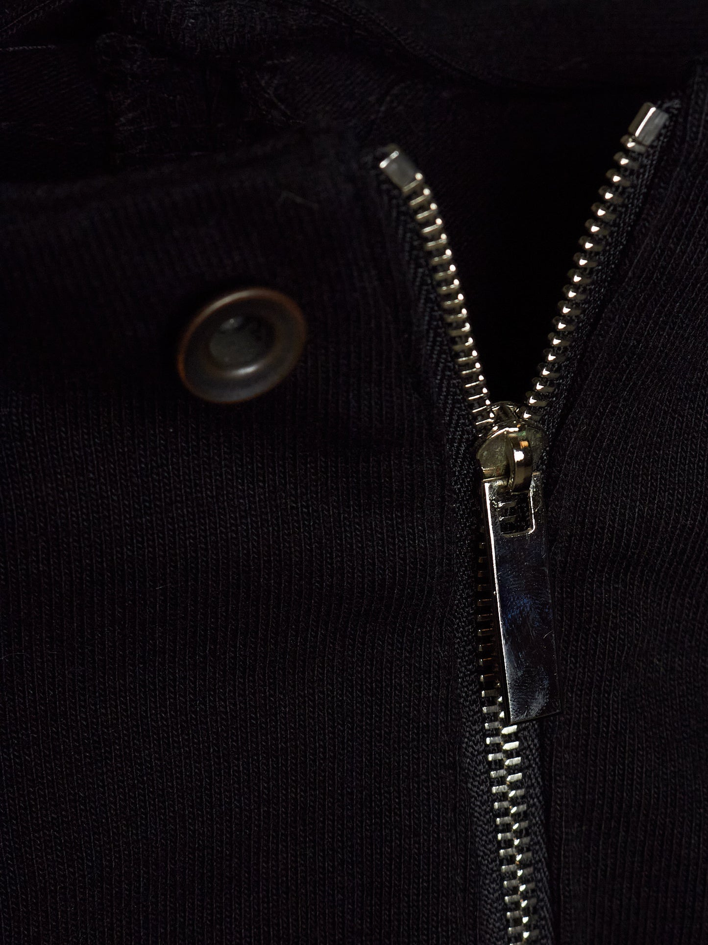 Close-up detail of a black ribbed knit garment featuring a silver metal zipper and a dark metal snap button.