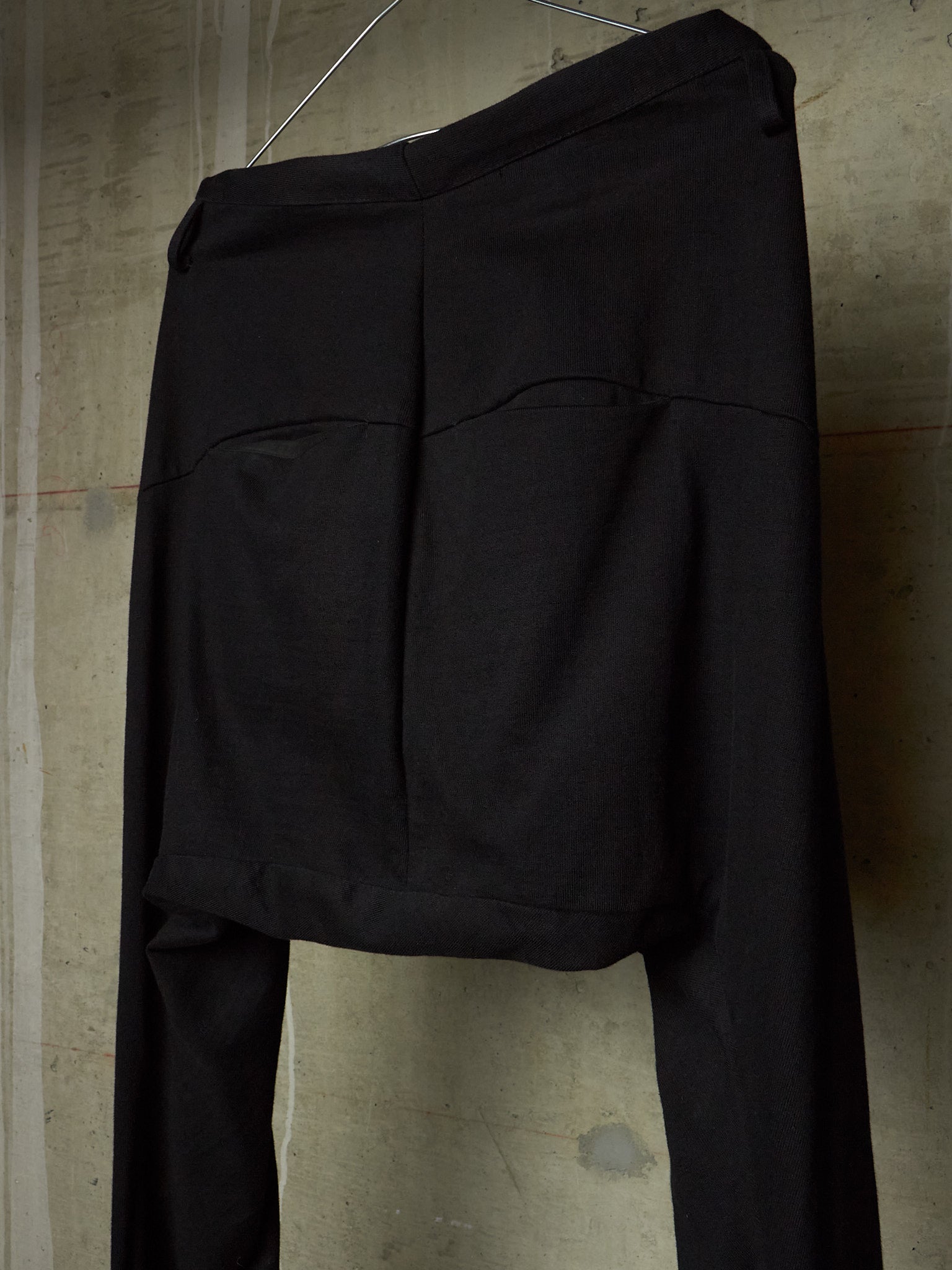 Black wide-leg trousers with a unique draped, asymmetrical front design, hanging against a textured concrete wall.