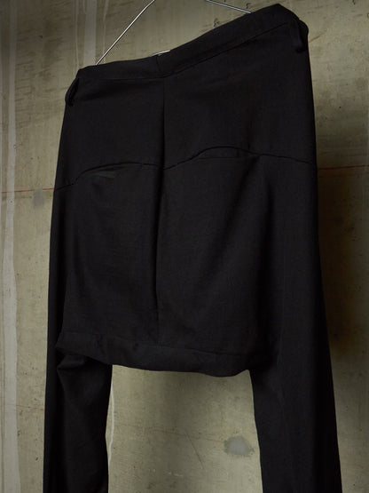 Black wide-leg trousers with a unique draped, asymmetrical front design, hanging against a textured concrete wall.