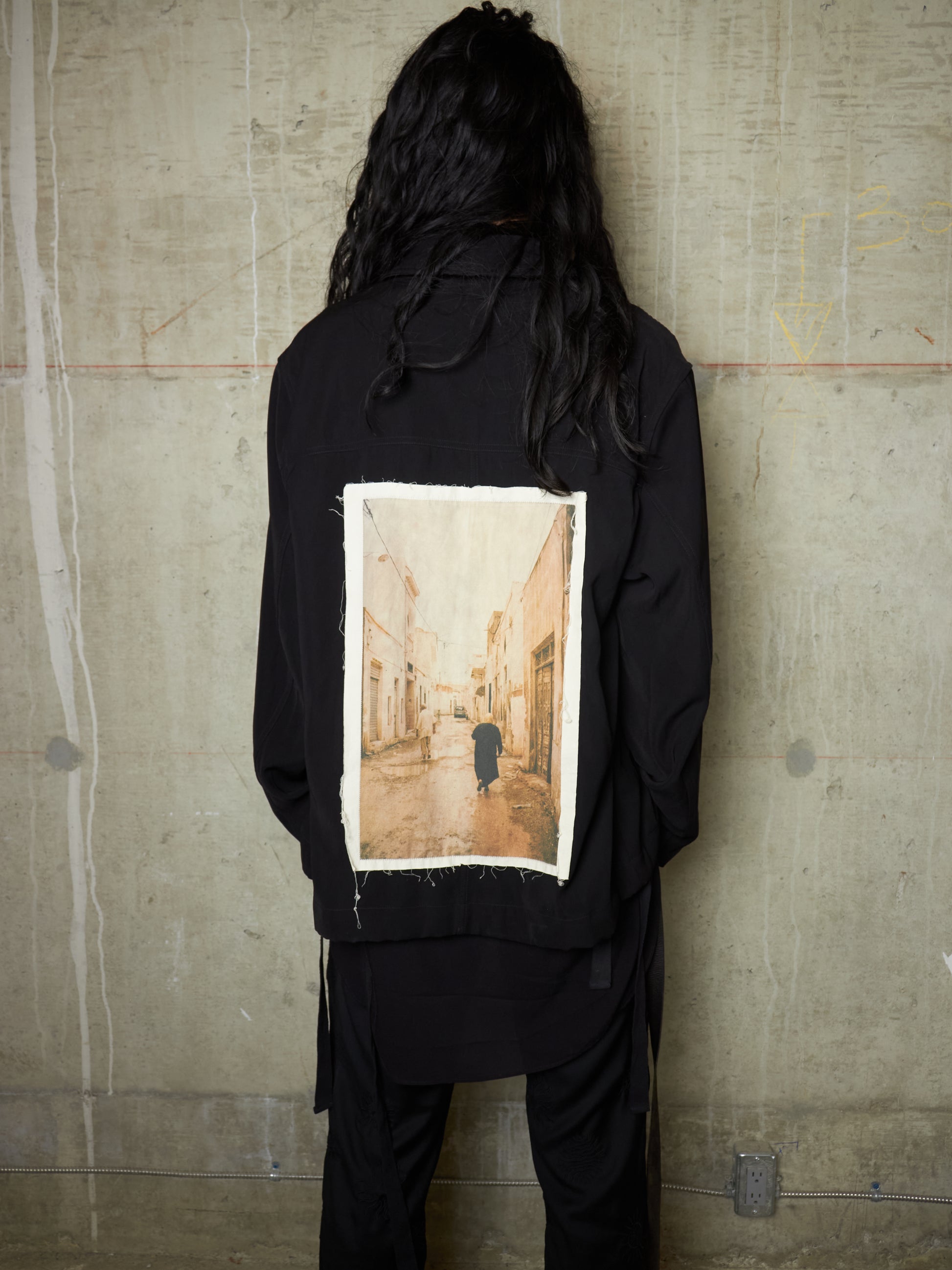 Person wearing a black jacket with a printed image on the back against a textured wall.
