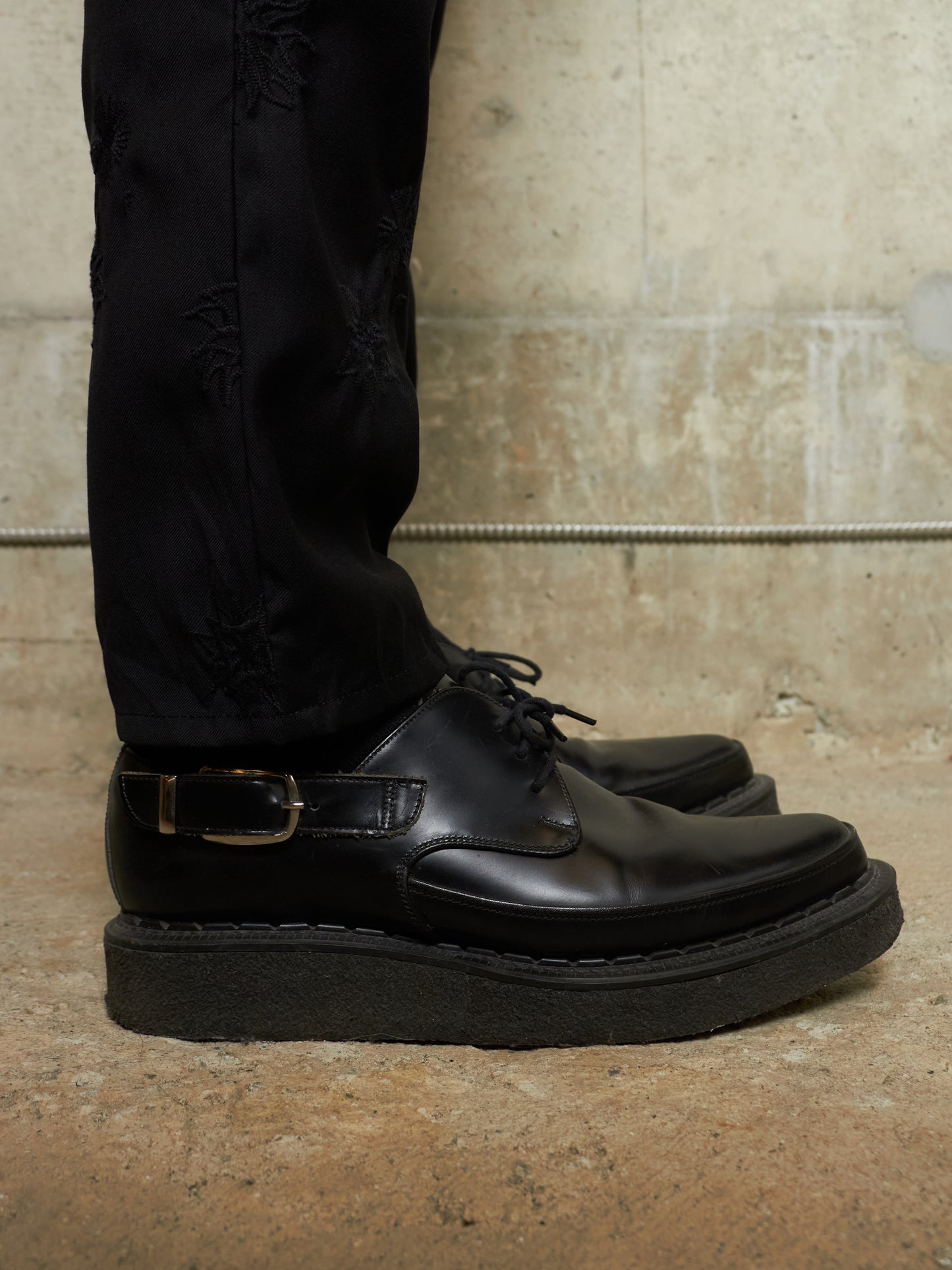 Black leather shoes worn with black pants against a textured wall.