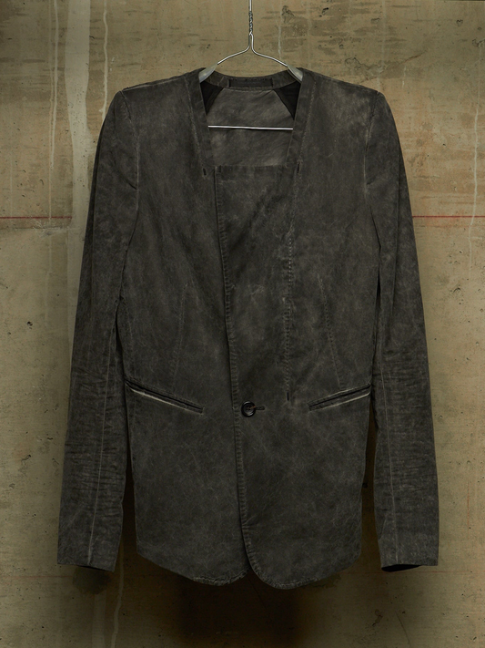 A dark charcoal gray distressed leather blazer jacket hanging on a metal hanger against a textured concrete wall.