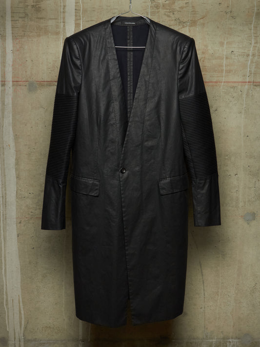 A black leather long coat with textured ribbed sleeves, hanging against a textured concrete wall.