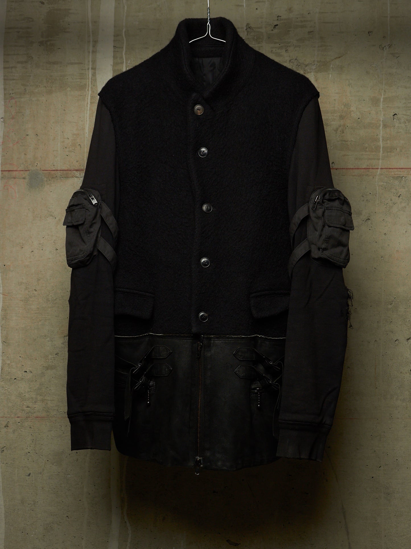A black asymmetrical jacket with a high collar, button front, and unique padded sleeve details, hanging against a textured concrete wall.