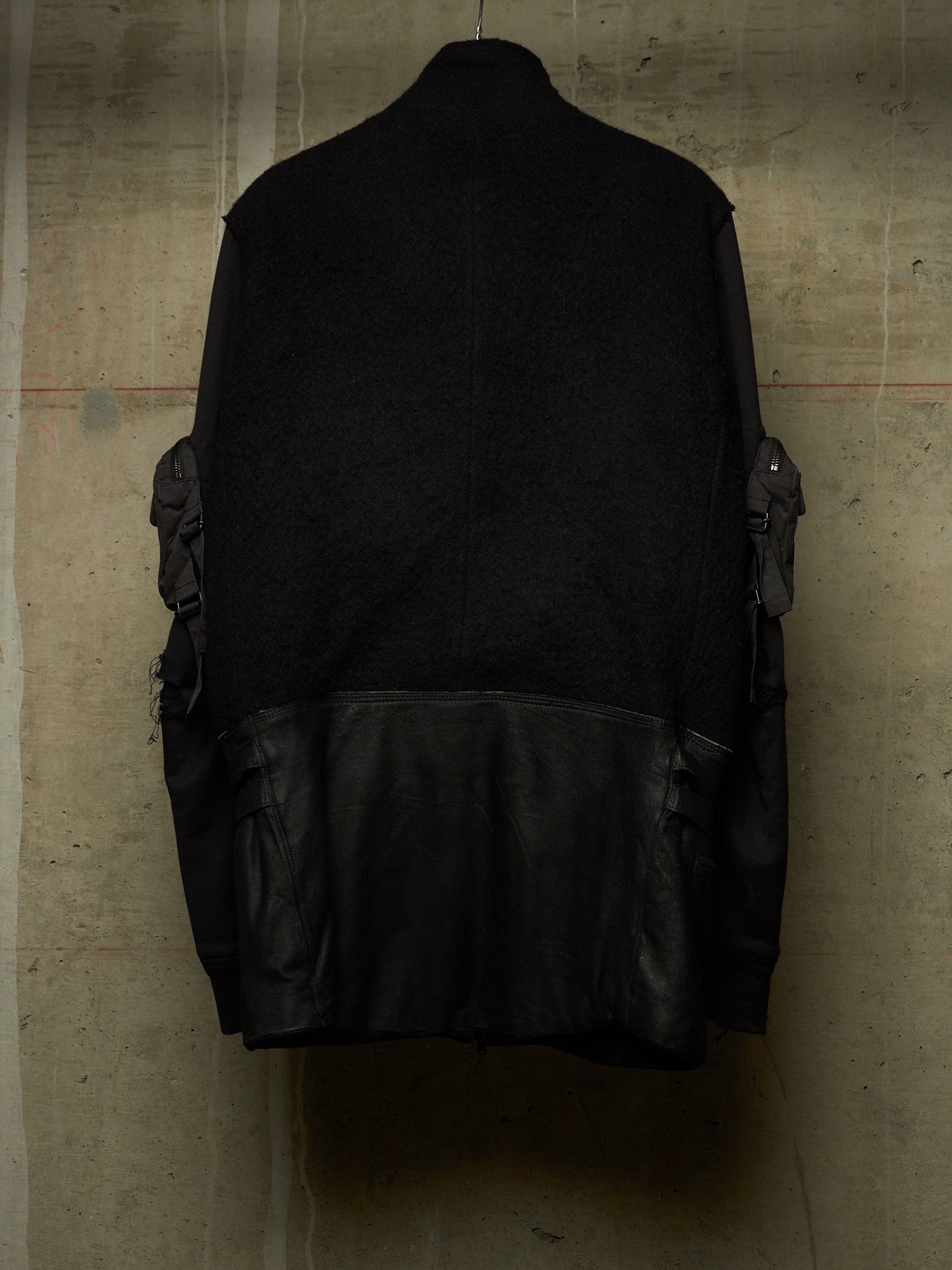 A black jacket with a textured knit upper body and smooth leather lower panel, shown hanging against a concrete wall.