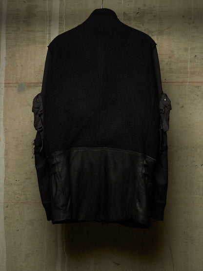 A black jacket with a textured knit upper body and smooth leather lower panel, shown hanging against a concrete wall.