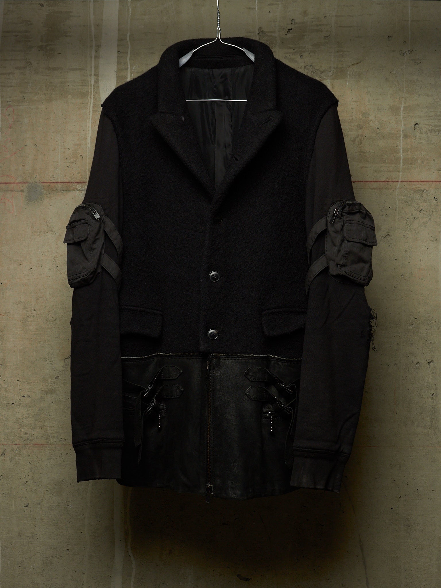 A black heavy-texture jacket with contrasting dark gray fabric on the sleeves and multiple utility pockets, hanging against a textured concrete wall.