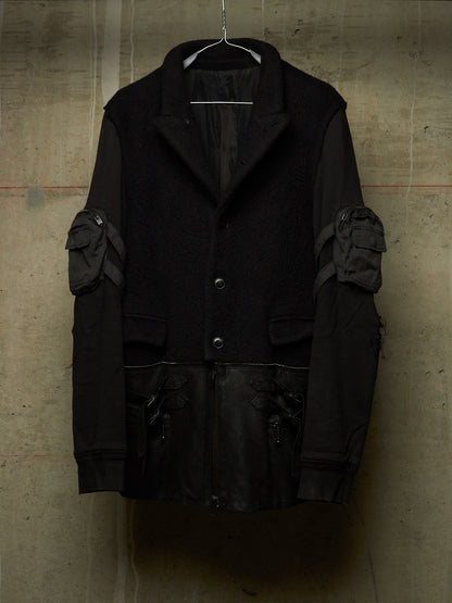 A black heavy-texture jacket with contrasting dark gray fabric on the sleeves and multiple utility pockets, hanging against a textured concrete wall.