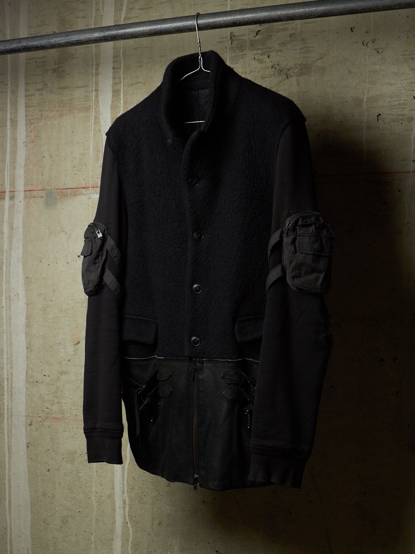 A black utility jacket with textured wool-like body panels and matte black leather sleeves, hanging on a metal hanger against a textured concrete wall.