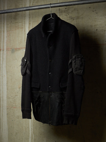 A black utility jacket with textured wool-like body panels and matte black leather sleeves, hanging on a metal hanger against a textured concrete wall.