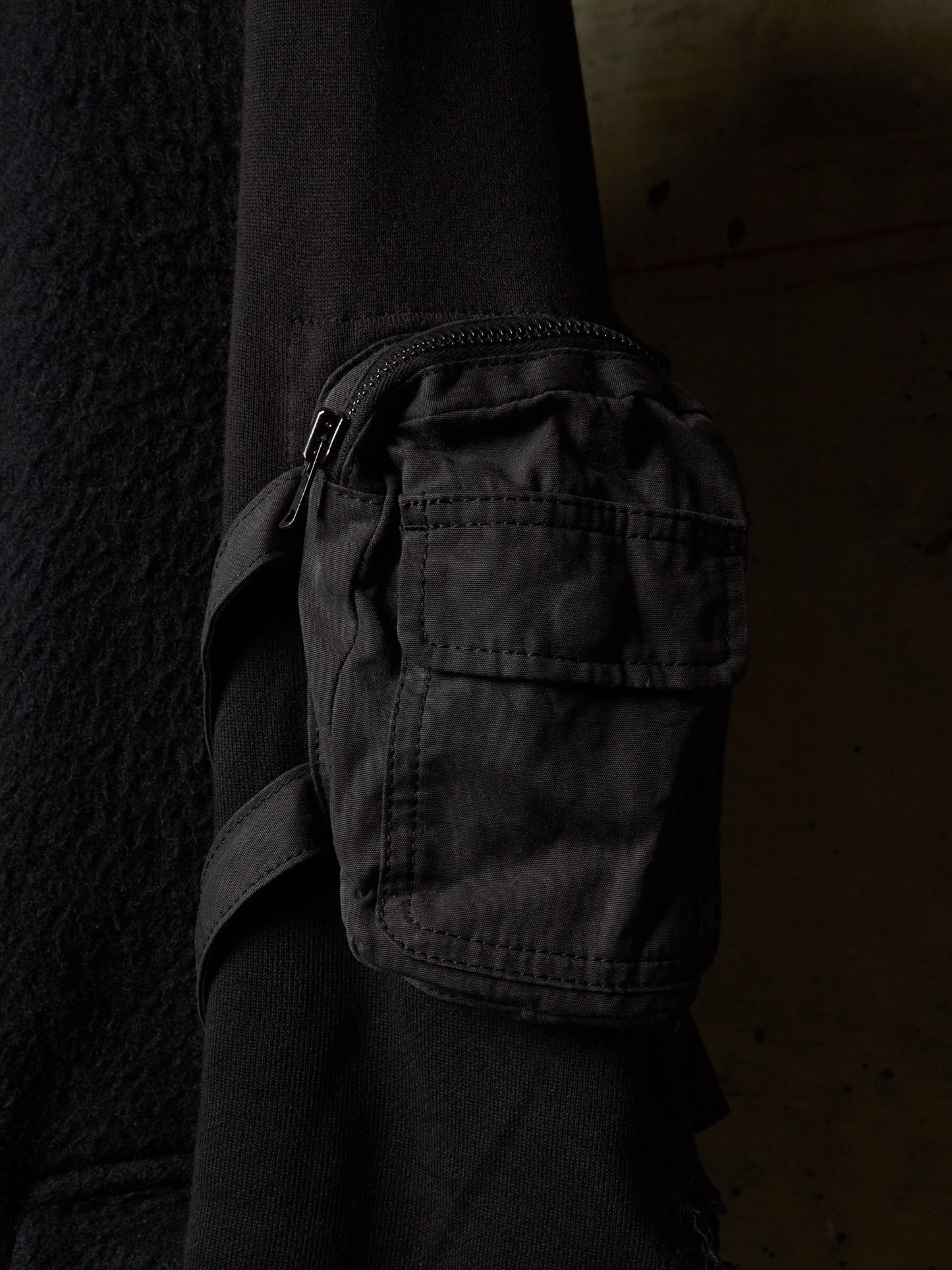 A close-up of a black utility pouch with a zipper and strap, attached to a black sleeve, set against a dark textured background.