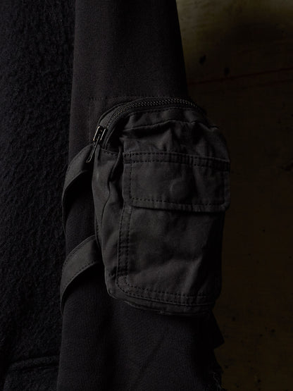 A close-up of a black utility pouch with a zipper and strap, attached to a black sleeve, set against a dark textured background.